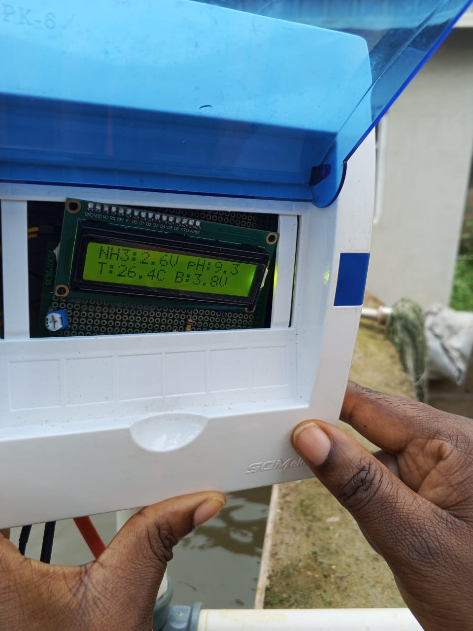 Hands holding an open white monitoring enclosure with a blue cover and an LCD showing water-quality readings beside a fish tank.